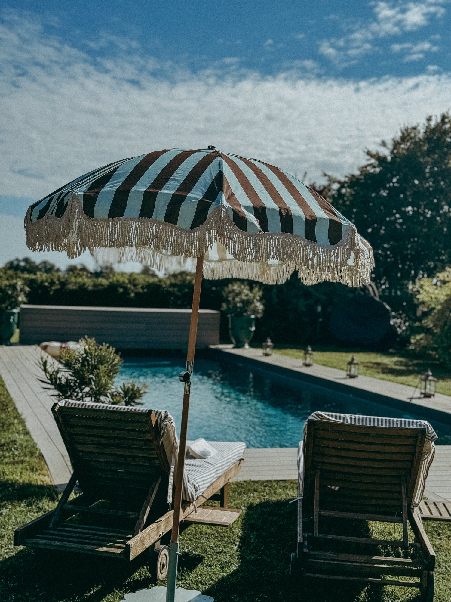 Mary blue and brown striped parasol with cotton fringing and beech wood frame. Pool setting, garden with pool and parasol on patio area.