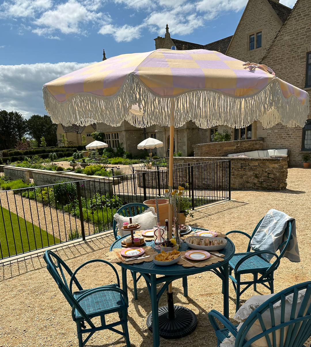 The Mabel boho chic parasol with lilac and cream checkerboard print, natural cotton fringing, adjustable wooden frame pole, collapsible design, and matching canvas carry bag.