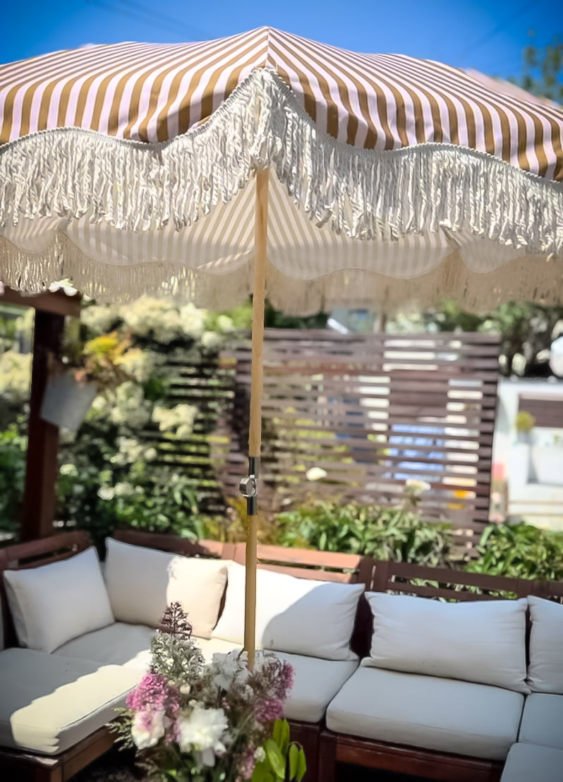 The Gigi luxury pink and camel striped parasol with natural cotton fringing, wooden frame pole, adjustable positioning, collapsible design, and matching carry bag.