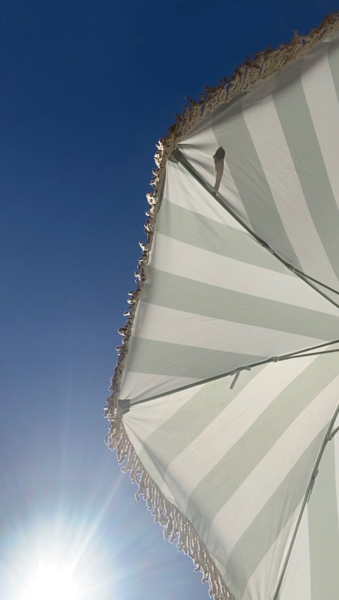 Sage green stripe parasol with natural cotton fringing, showcasing the Clara design in a stylish outdoor setting.