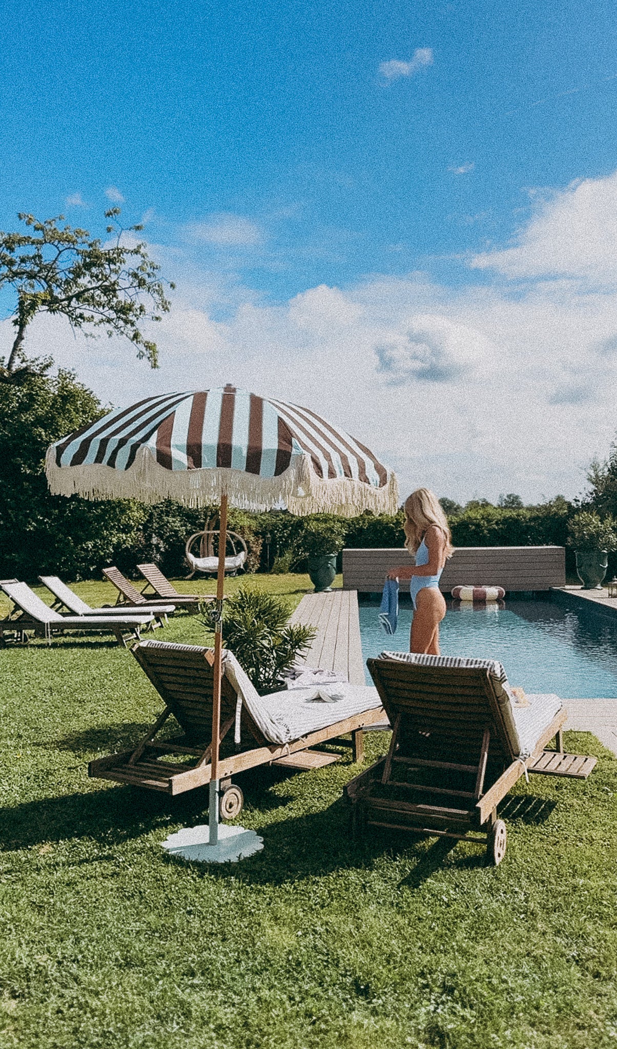 Mary blue and brown striped parasol with cotton fringing and beech wood frame. Pool setting, garden with pool and parasol on patio area.