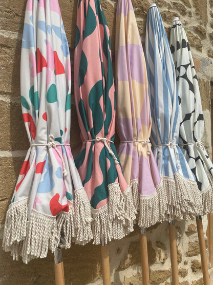 The Mabel boho chic parasol with lilac and cream checkerboard print, natural cotton fringing, adjustable wooden frame pole, collapsible design, and matching canvas carry bag.