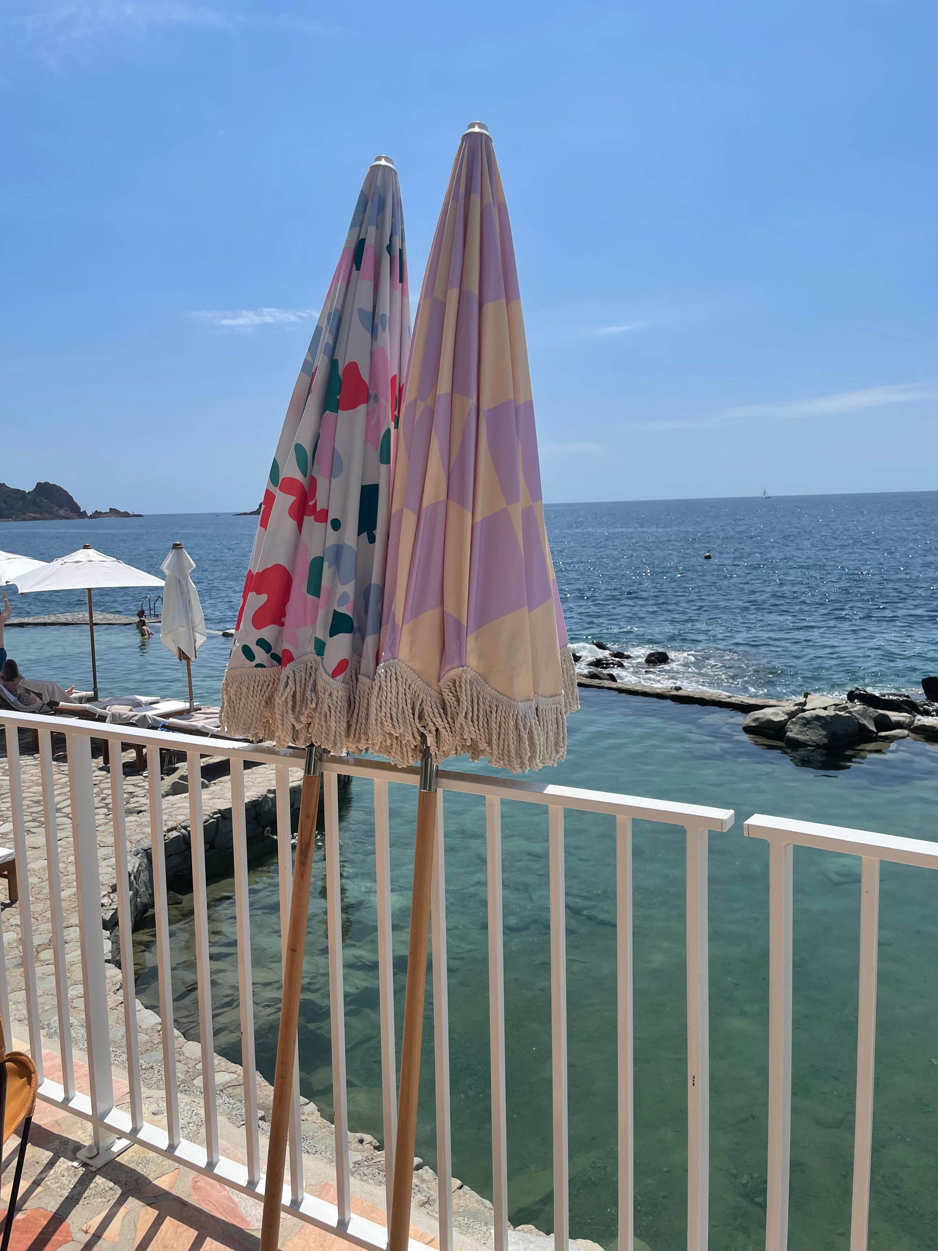 Mabel checkerboard lilac and cream parasol with natural cotton fringing, positioned by the poolside for a stylish outdoor setting.